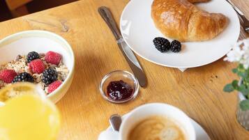 Un desayuno con un croissant, bayas, un tazón de cereal, un recipiente pequeño de mermelada y una taza de café sobre una mesa de madera.