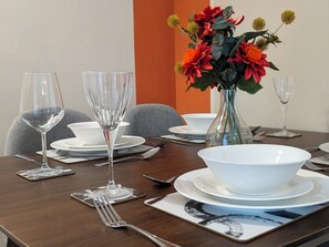 A dining table set with white plates, bowls, cutlery, and two wine glasses. A vase with red and orange flowers is placed on the table. - Salford | FreePPrking | WasherDryer | GroundFloor (Manchester)