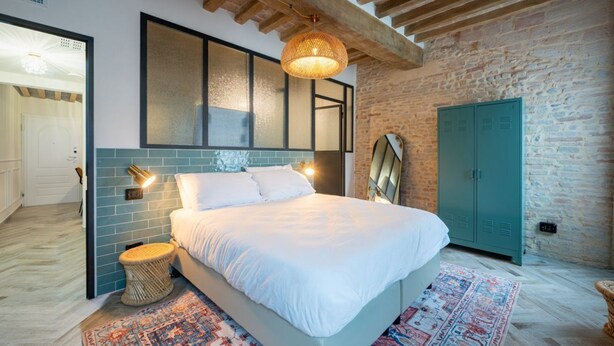 A bedroom with a large bed, a woven stool, a patterned rug, a green wardrobe, and a stone wall.