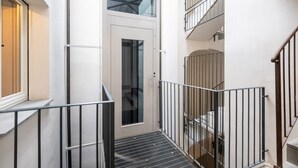 A narrow indoor corridor with a metal railing, a door with a glass panel, and a staircase with a metal railing.