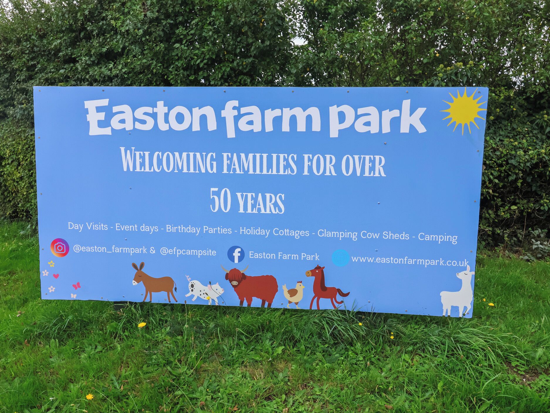 Een blauw bord van Easton Farm Park, dat gezinnen al meer dan 50 jaar verwelkomt, met activiteiten zoals dagbezoeken, evenementen, verjaardagsfeesten, vakantiehuisjes, kamperen in luxe tenten, koeienstallen en een camping.