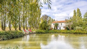 Uno stagno tranquillo con una casa sullo sfondo, immerso nel verde e circondato da alberi alti.