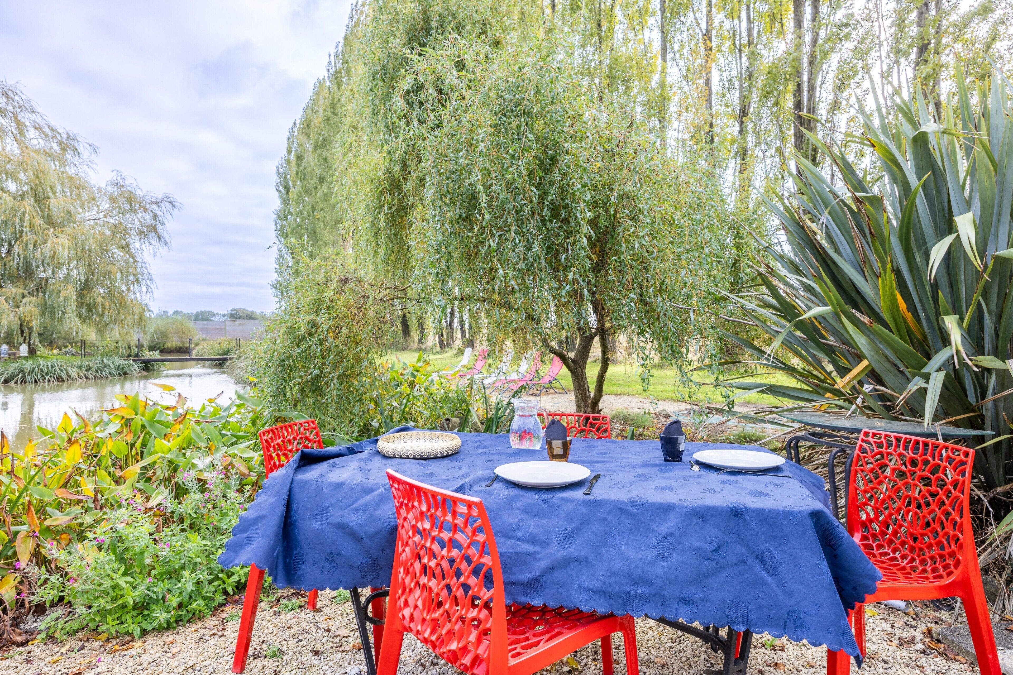Une table dressée à l’extérieur, recouverte d’une nappe bleue, avec des chaises rouges, et offrant une vue sur un étang et des arbres.