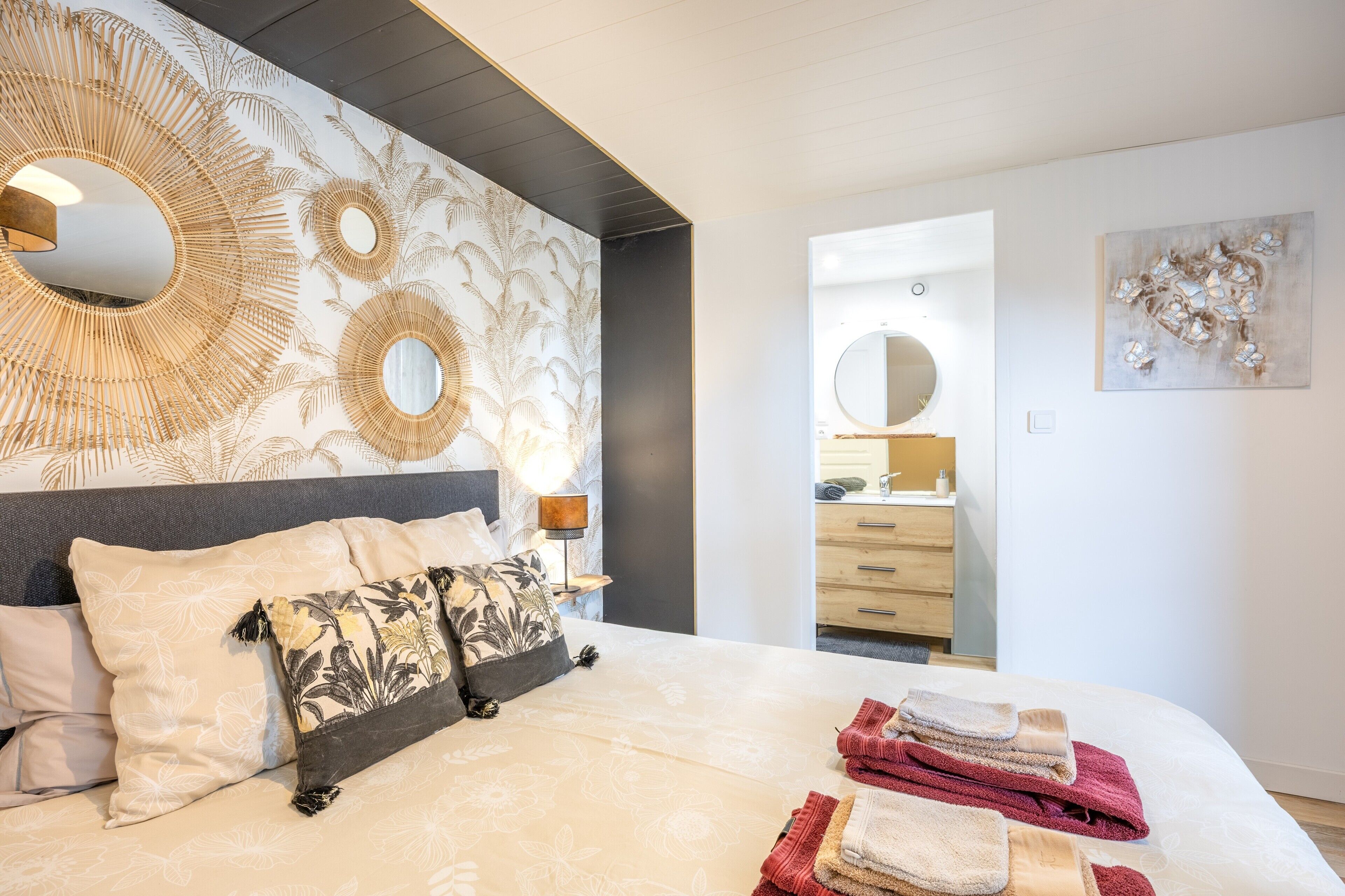A bedroom with a bed, pillows, a nightstand, a lamp, a mirror, a dresser, and towels.