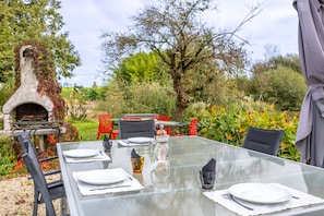 Un espace extérieur aménagé pour les repas, avec une table en verre, des chaises en métal et un four en pierre.