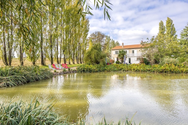 A serene pond with a house in the background, surrounded by lush greenery and tall trees.