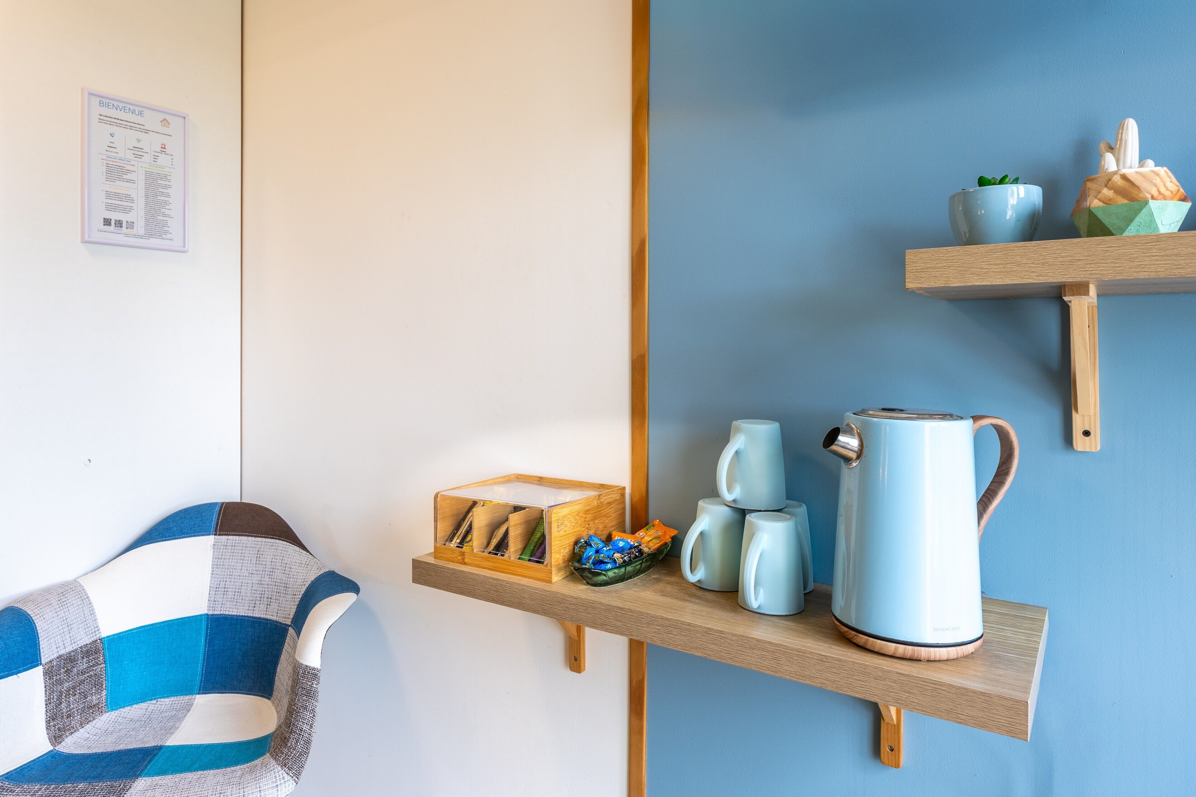 A room with a checkered chair, a wooden shelf with mugs, a kettle, and a small plant.