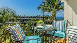 A balcony with two chairs and a table, overlooking a beach with palm trees and a clear sky. - New Listing! Akahi C613 – Ocean View Top Floor Condo, Newly Updated, Sleeps 4 (Kihei)