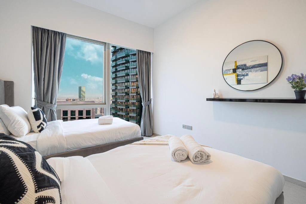 Ein modernes Hotelzimmer mit zwei Betten, einem großen Fenster mit Vorhängen, Blick auf die Stadt, einem runden Spiegel und einer kleinen Ablagefläche mit einer Pflanze.