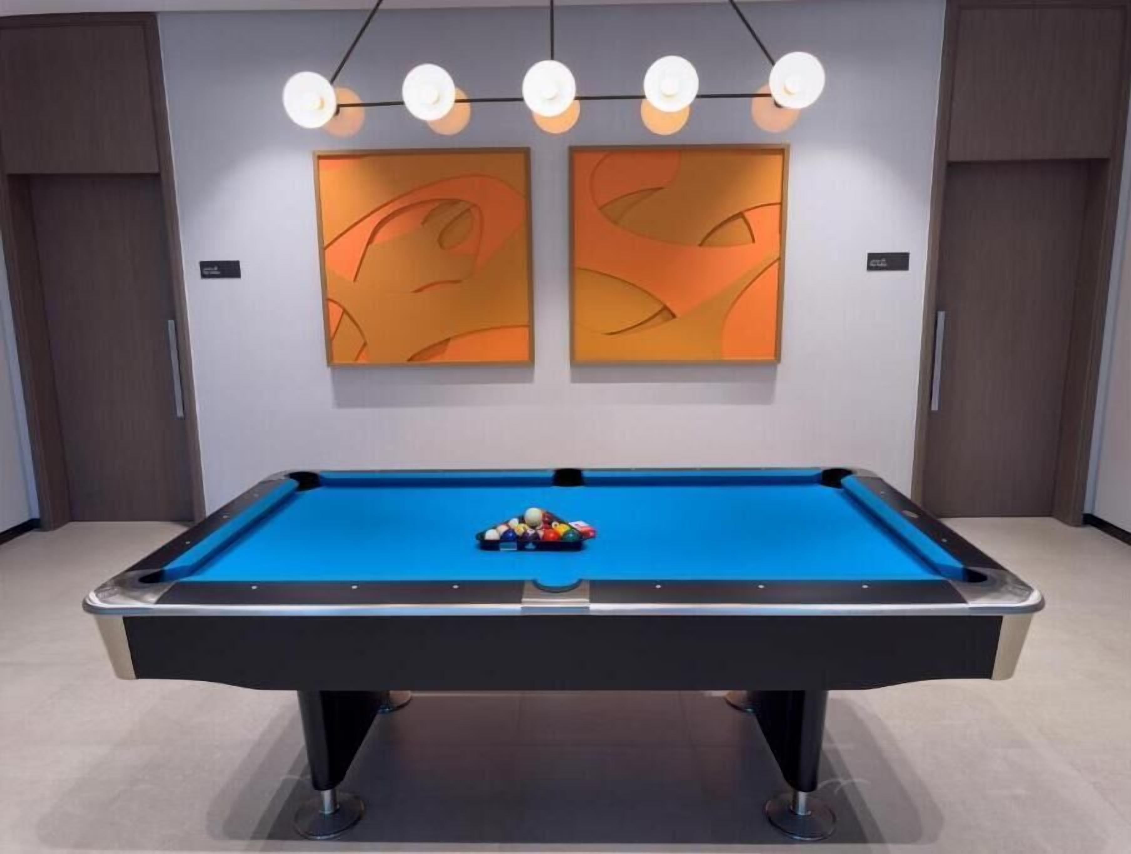 A pool table with a blue felt surface, surrounded by a modern room with two abstract paintings on the wall and a string of lights above.