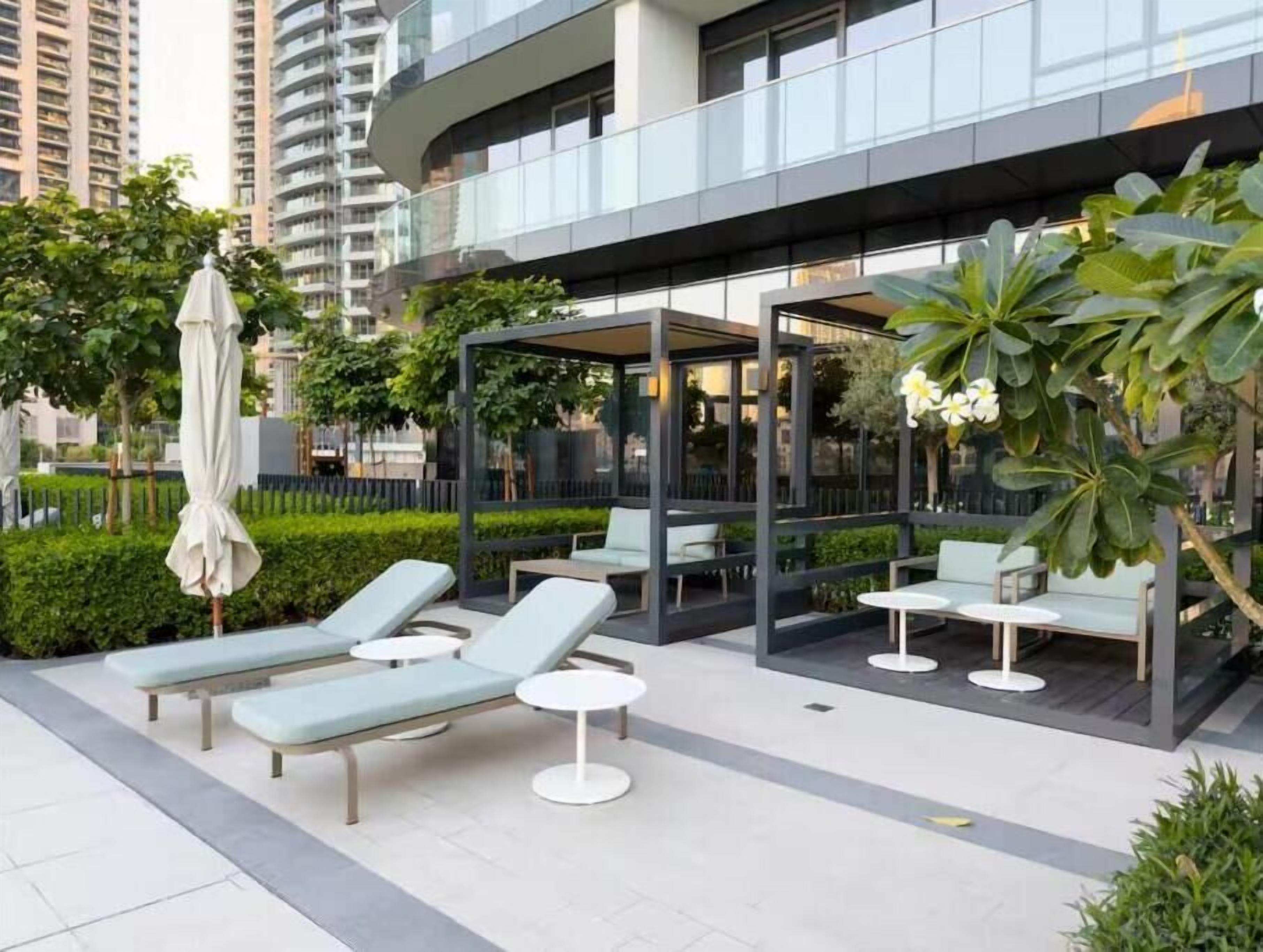An outdoor lounge area with modern seating, including lounge chairs and small tables, surrounded by greenery and a glass building in the background.