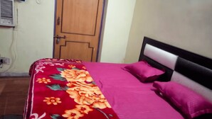 A bed with a floral bedspread and two pillows, a wooden door, and a wall-mounted air conditioning unit. - Hotel Raj Ratan (Kanpur)