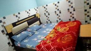 A single bed with a floral bedspread and a red blanket with a geometric pattern. The headboard is black with a gold accent, and the wall behind the bed has a checkered tile design. - Hotel Raj Ratan (Kanpur)