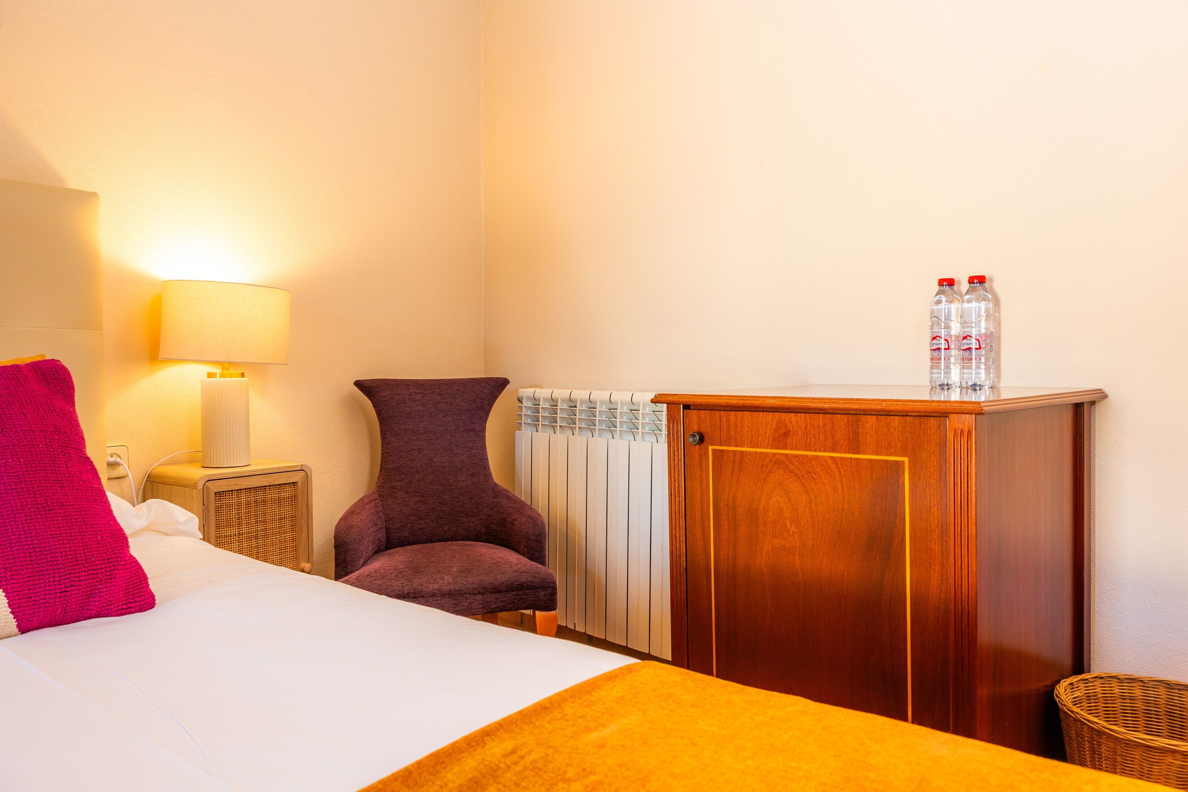 A hotel room with a bed, a lamp, a chair, a radiator, and a wooden cabinet with water bottles on top.
