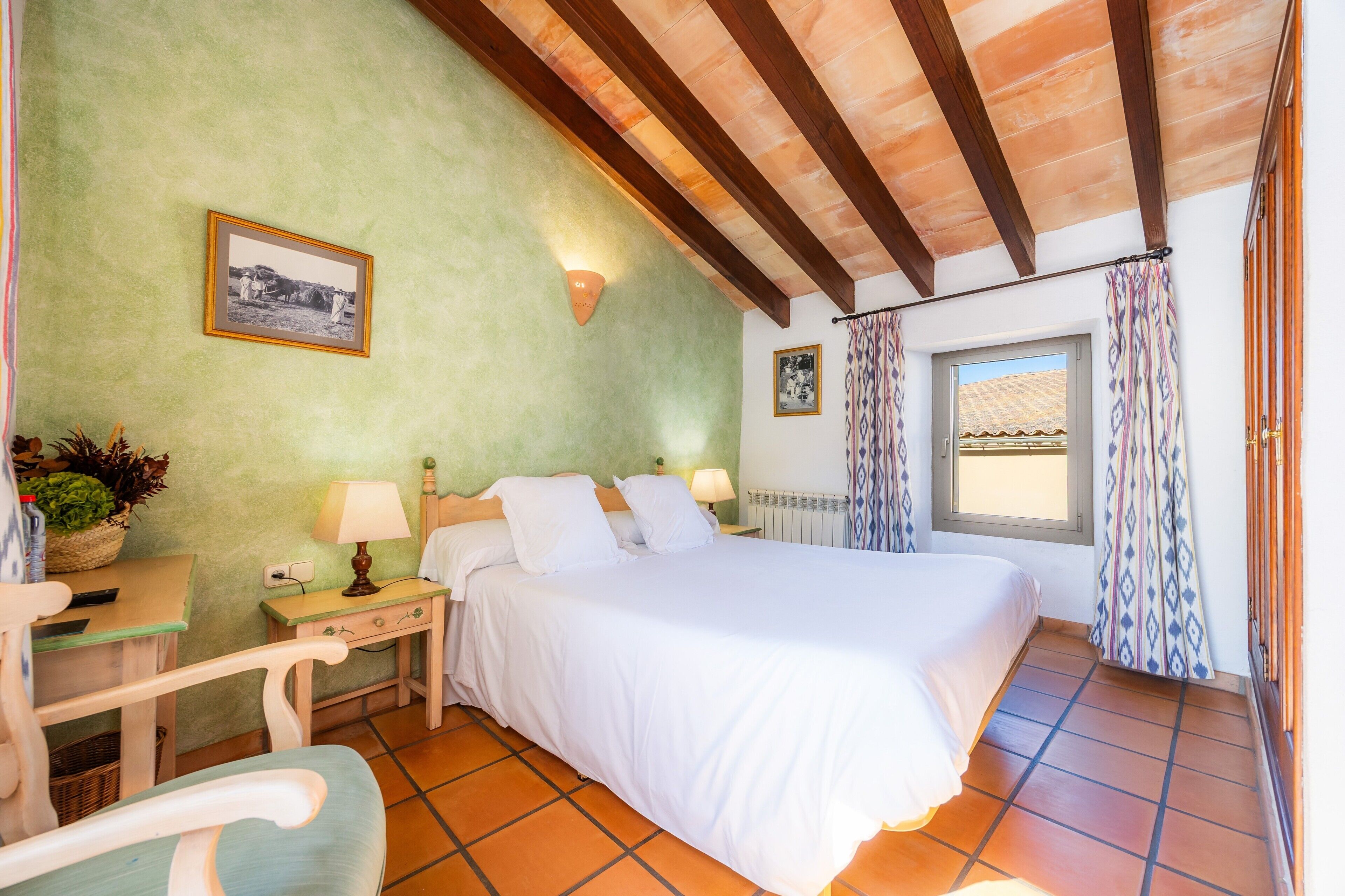 Un dormitorio con cama, mesitas de noche, lámpara, un cuadro en la pared y una ventana con cortinas.