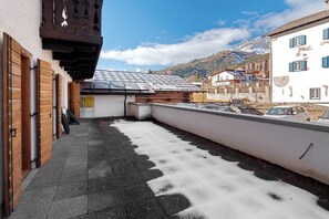 A rooftop terrace with a view of snow-capped mountains, a wooden pergola, and a white building with blue shutters.