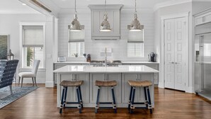 A modern kitchen with white cabinetry, a large island with stools, pendant lighting, and a stainless steel refrigerator.
