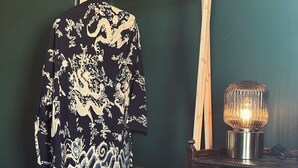 A room with a dark green wall, a wooden easel holding a patterned garment, a framed drawing, a woven basket, and a table with glasses and a lamp. - Margate Manor (Margate)