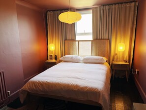 Una cama bien tendida con sábanas blancas, dos mesitas de noche con lámparas y una ventana con cortinas.