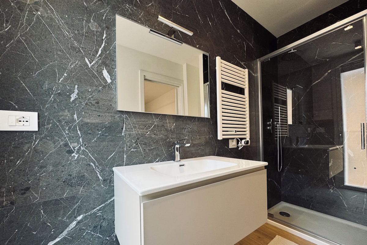 A modern bathroom with a white vanity, a large mirror, a black marble wall, and a glass shower enclosure.