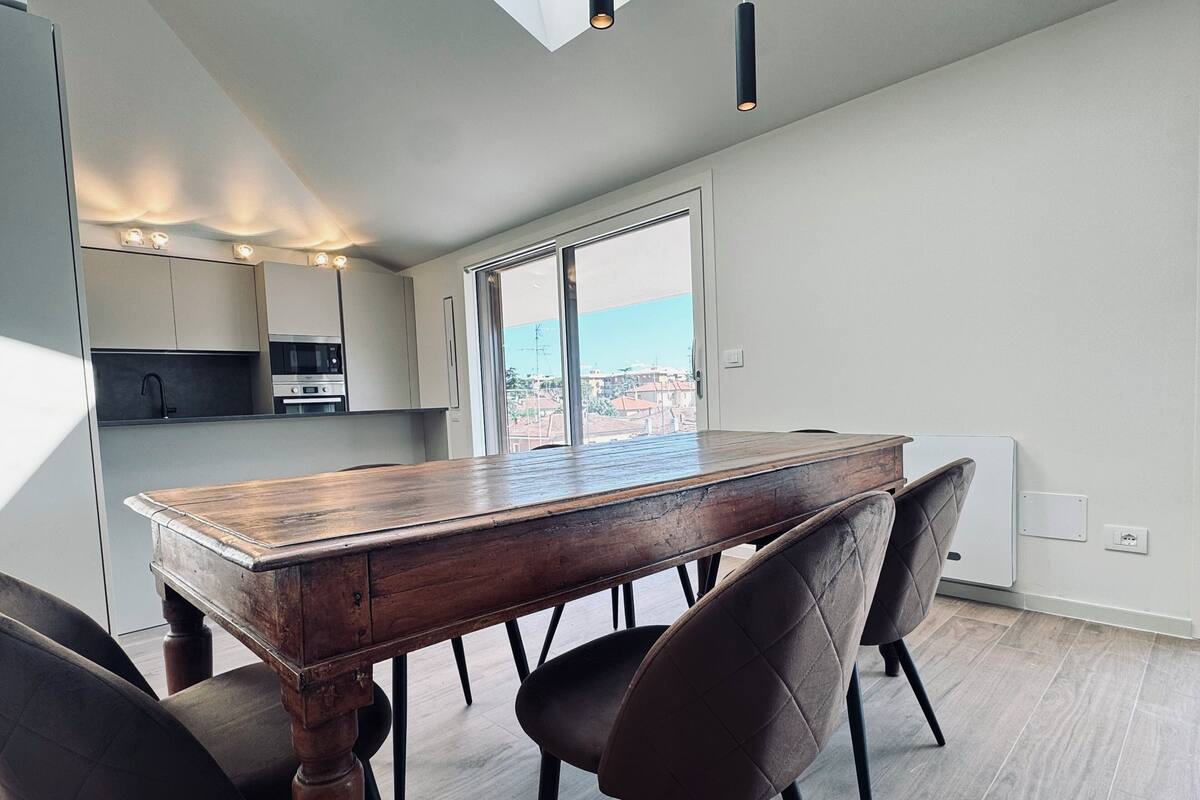 A modern kitchen with a large wooden table and four chairs, a window with a city view, and a built-in oven.