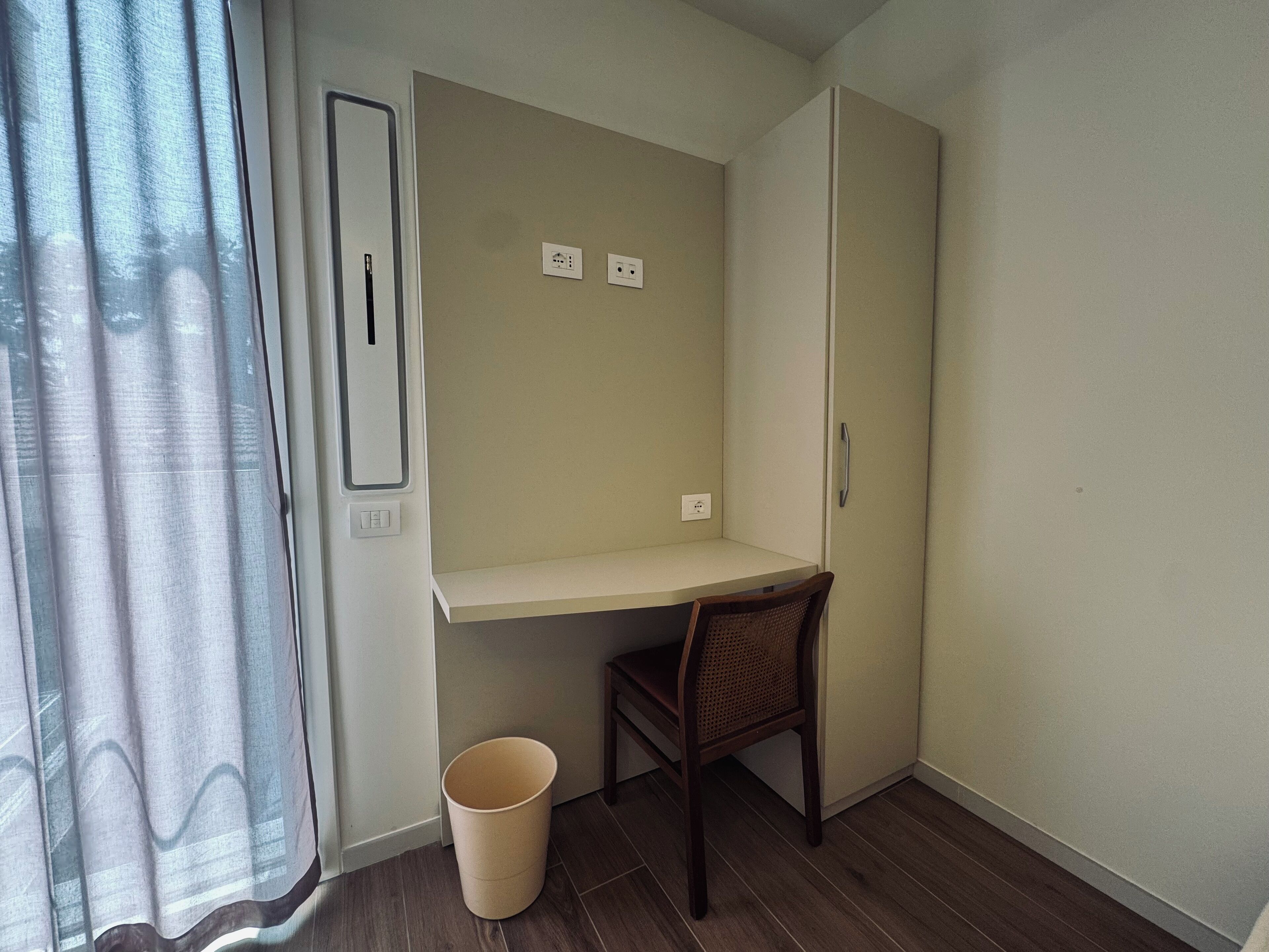 A small room with a wooden desk, a chair, and a trash bin. There are two electrical outlets on the wall.