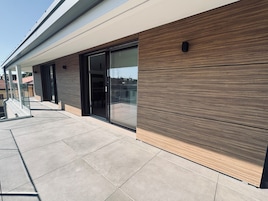 A modern building exterior with a glass door entrance, a covered outdoor seating area, and a tiled patio.