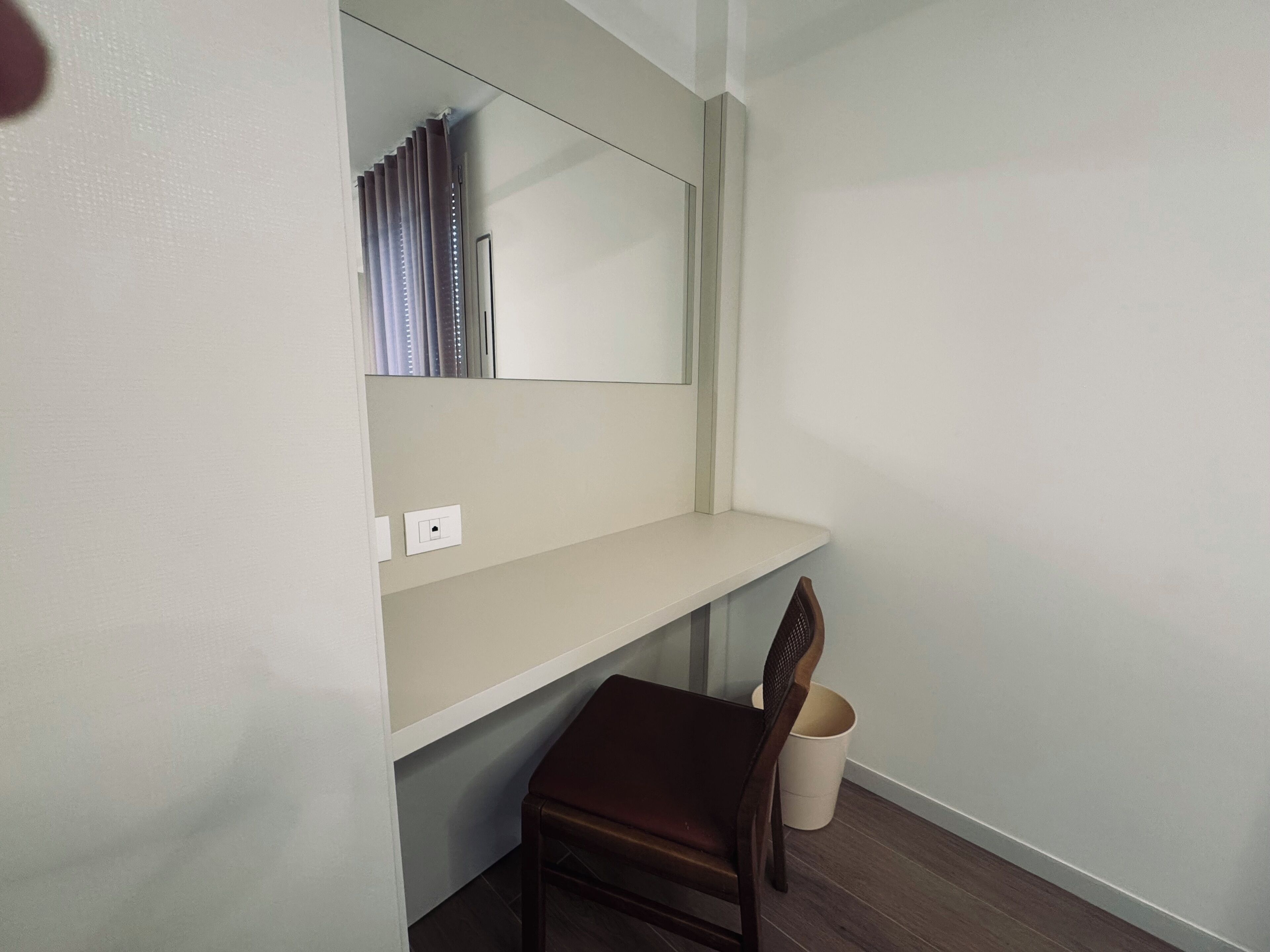 Una stanza piccola e pulita con una sedia di legno, uno scaffale bianco e uno specchio appeso al muro.