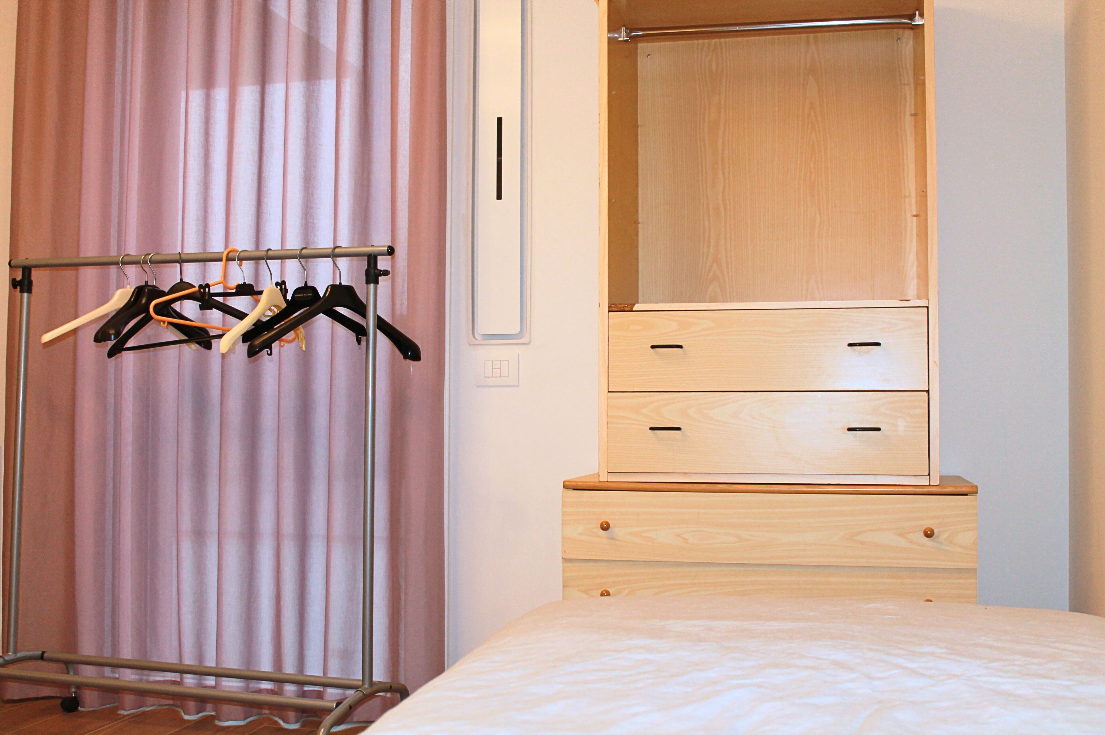 Quarto com cama, cómoda de madeira com gavetas, guarda-roupa com espaço para pendurar e suporte de metal com cabides.