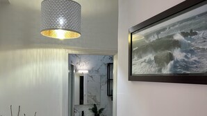 Een gang met een marmeren muur, een lijst met een schilderij van een zeegezicht, een hanglamp en een potplant.