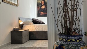 Een slaapkamer met een bed, nachtkastje, lamp en een groot schilderij van de Mona Lisa aan de muur.