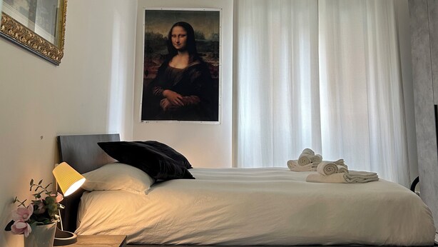 Een slaapkamer met een bed, een plafondventilator, een schilderij van de Mona Lisa, een nachtkastje en een raam met gordijnen.