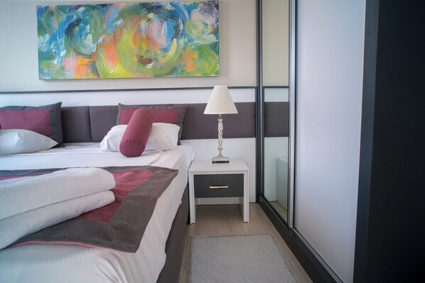Un dormitorio moderno con una cama grande, una mesita de noche con lámpara, un cuadro abstracto de colores en la pared y vistas al exterior a través de un ventanal.