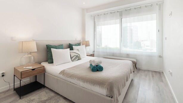 Un dormitorio moderno con una cama grande, mesitas de noche, una ventana con cortinas y vistas a la ciudad.