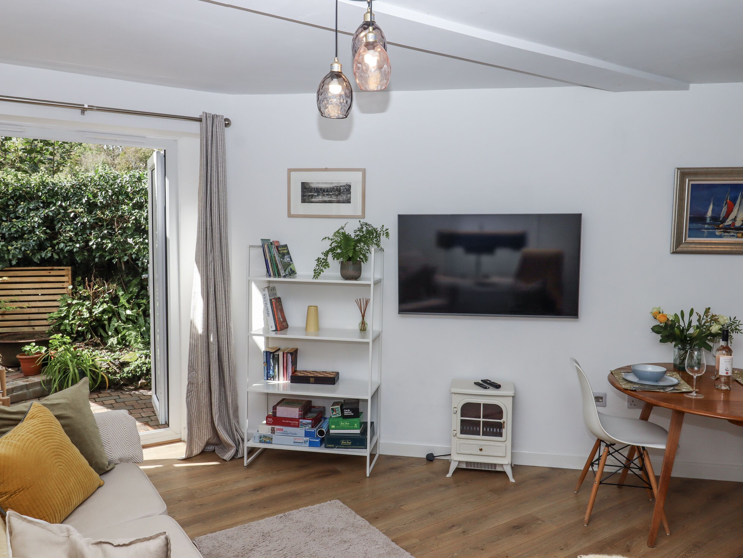A modern living room with a white bookshelf, a small white stove, a dining area with a wooden table and chairs, and a large window overlooking greenery.