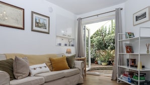 Ein Wohnzimmer mit einem beigefarbenen Sofa, einem Couchtisch und einem Bücherregal. Eine Tür führt zu einem Garten mit Kübelpflanzen.