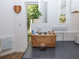 Una cocina con una cesta de mimbre que contiene un tarro, un bol y pantuflas, una planta en maceta y un adorno de madera con forma de corazón en la pared.