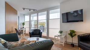 Una sala de estar moderna con un sofá, una mesa de centro, un televisor montado en la pared y ventanas amplias.