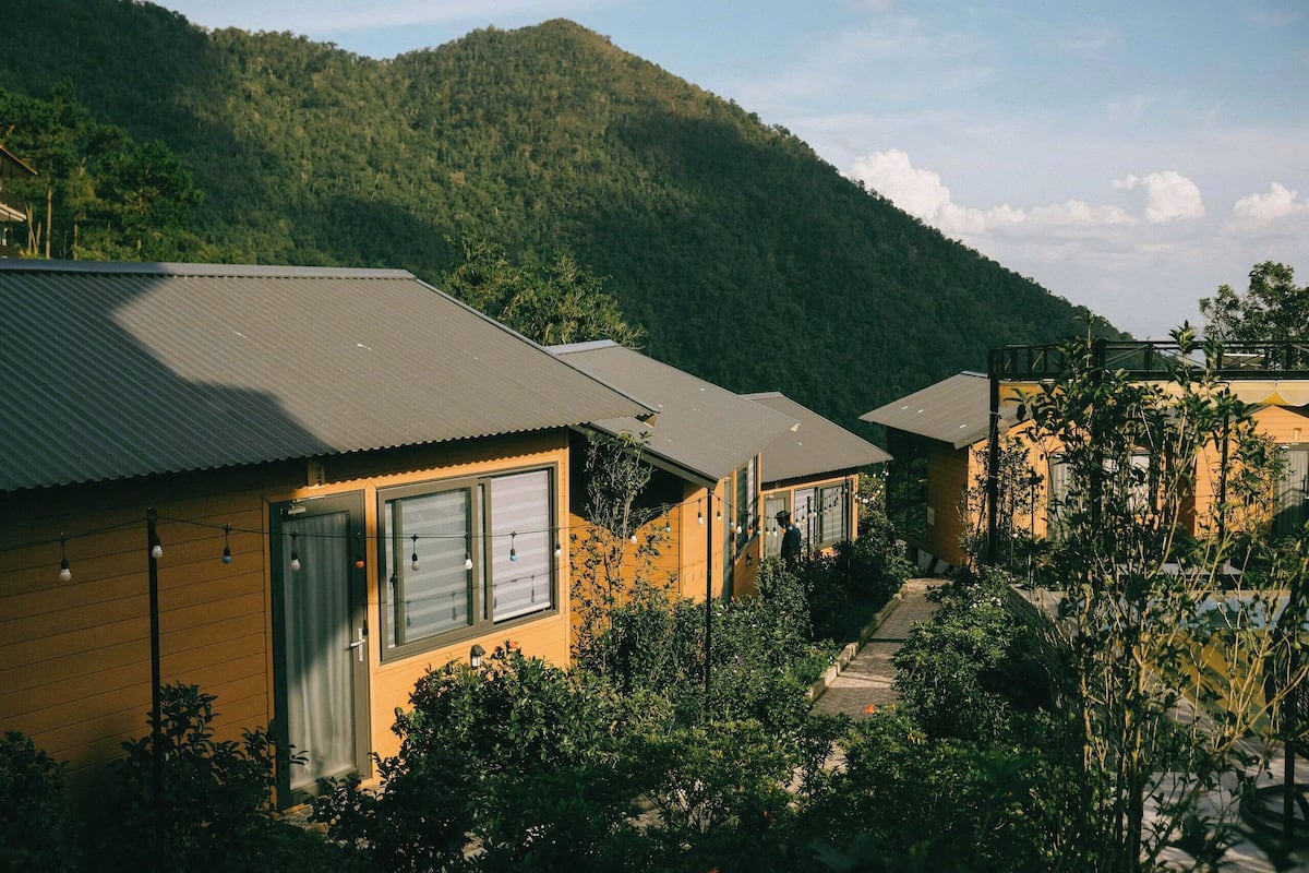 A row of cabins with a mountain backdrop, surrounded by greenery.