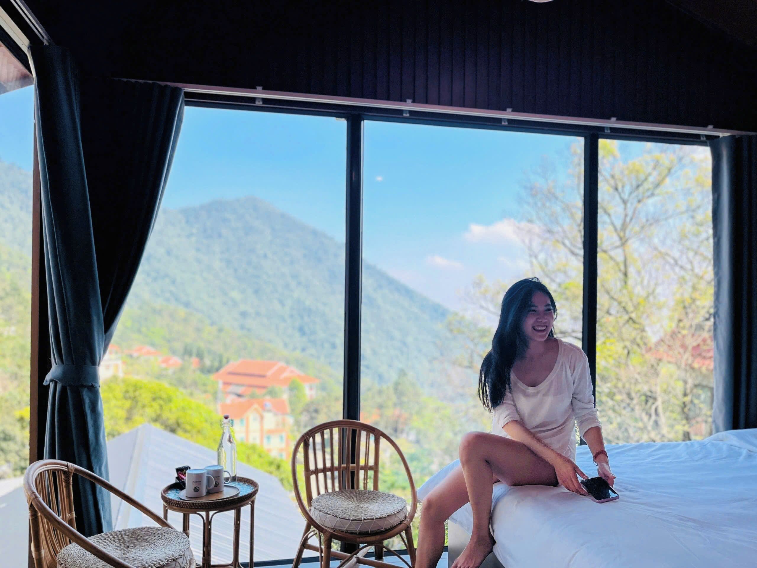 A room with a bed, a wicker chair, a small round table with a bottle and two mugs, and a view of mountains and greenery.