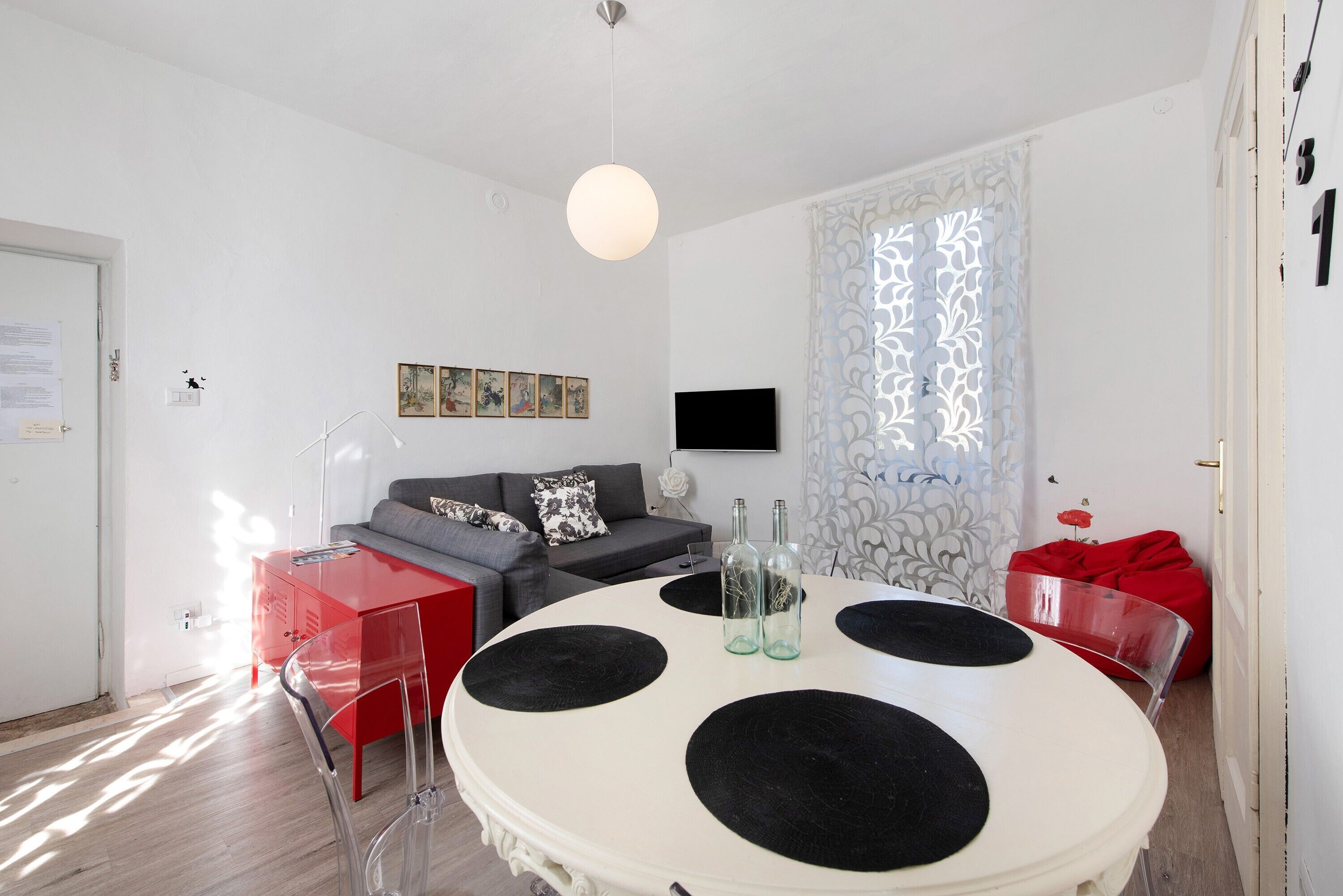 Ein modernes Wohnzimmer mit einem runden weißen Tisch, roten und transparenten Stühlen, einem grauen Sofa und einem roten Sitzsack.