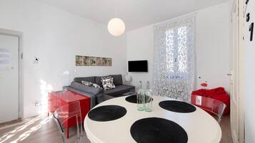 Ein modernes Wohnzimmer mit einem runden weißen Tisch, roten und transparenten Stühlen, einem grauen Sofa und einem roten Sitzsack.
