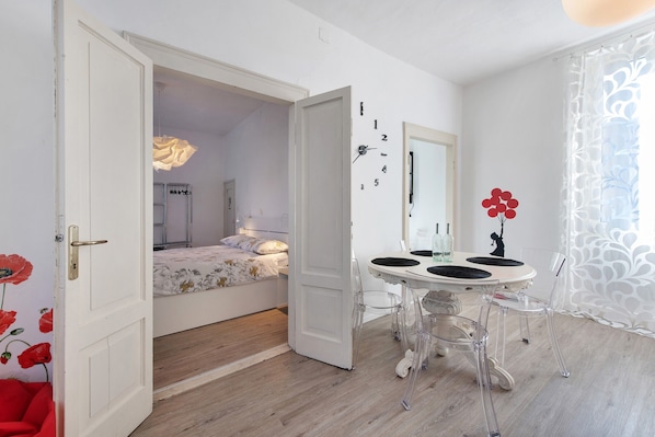 A room with a bed, a dining table with chairs, a wall clock, and a door leading to another room.