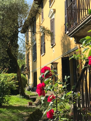 A yellow building with black shutters, a balcony, and a garden with red flowers.