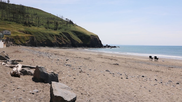 Ein Strand mit felsigem Vordergrund, eine grasbewachsene Anhöhe mit Bäumen und einem klaren Himmel.