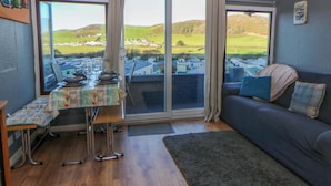 Ein gemütliches Zimmer mit Esstisch für zwei Personen, einer Couch mit Kissen und großen Fenstern mit Blick auf eine Wohngegend.