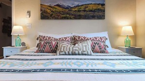 Una cama con almohadones estampados, un cuadro de paisaje en la pared y dos lámparas de noche.