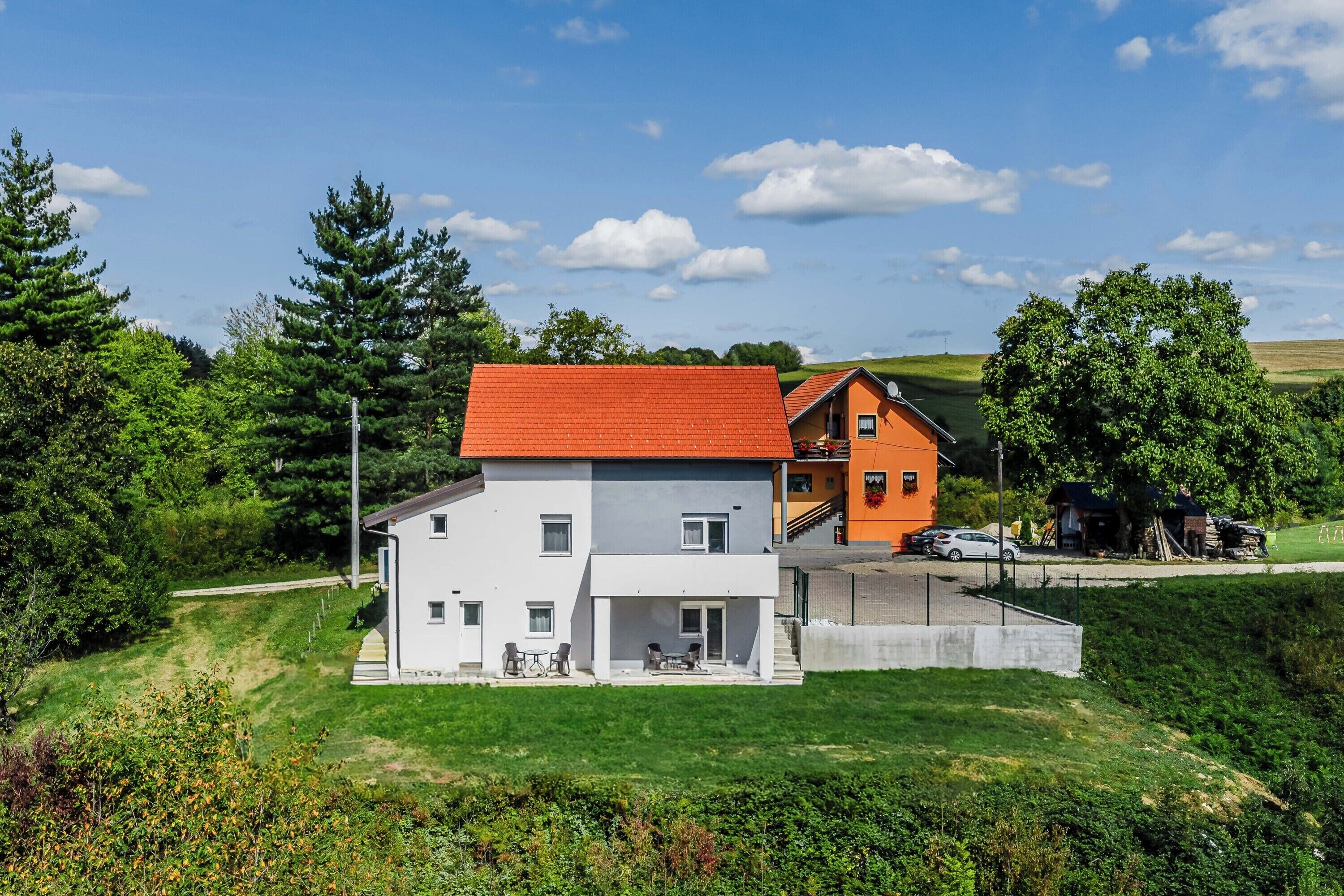 A two-story house with a red roof and white walls, surrounded by greenery and a clear sky.