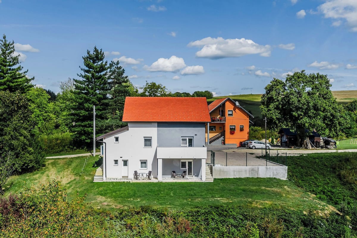 A two-story house with a red roof and white walls, surrounded by greenery and a clear sky.