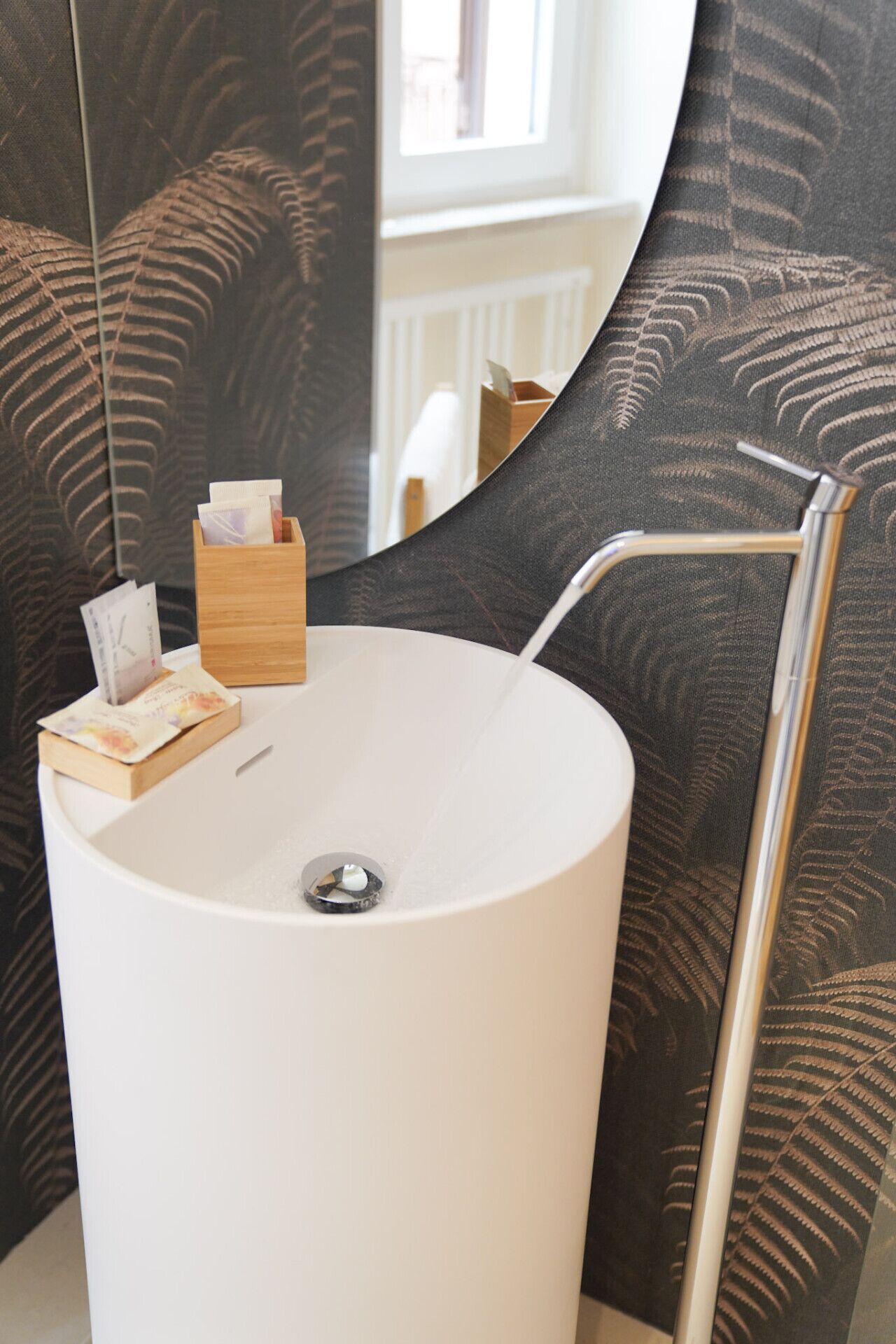Ein Badezimmer mit einem runden, weißen Waschbecken, einer Holztischbox für Taschentücher und einem Spiegel mit Farnmuster.
